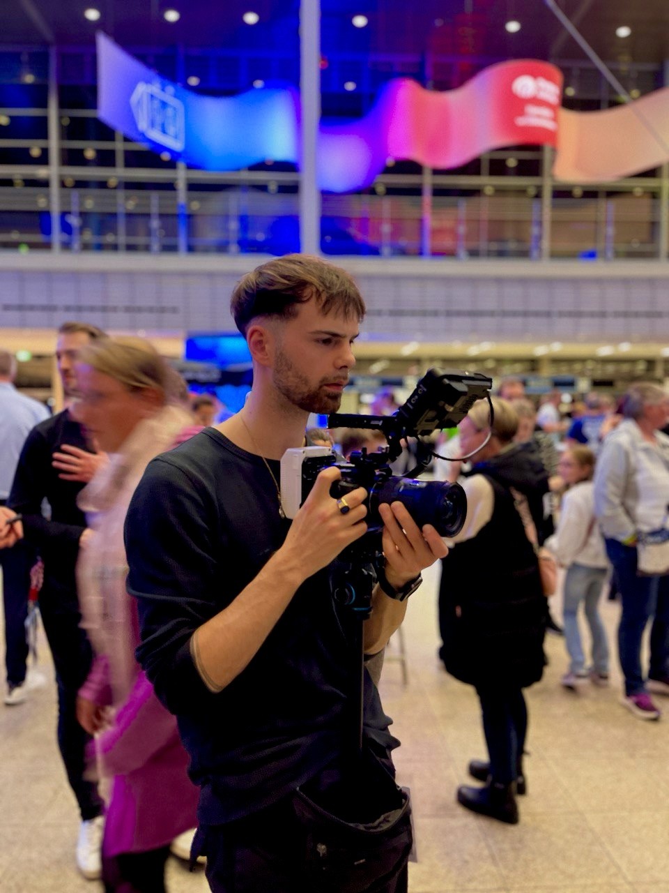 Ein professioneller Content Creator bei der Arbeit: Ein junger Mann in schwarzem Langarmshirt hält eine hochwertige Kamera mit externem Monitor, während er bei einem Event in einer modernen, hell beleuchteten Halle filmt. Im Hintergrund sind unscharfe Menschen und bunte Lichtinstallationen in Blau- und Rottönen zu sehen. Perfektes Bild für Themen wie Videoproduktion, Content Creation, Event-Dokumentation oder kreative Medienarbeit.