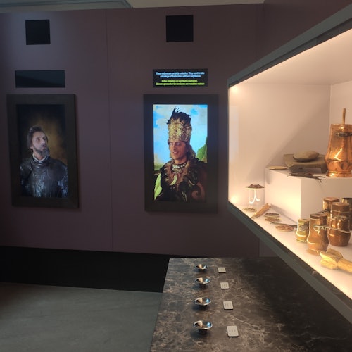 Muzejní výstava s portréty muže v brnění a osoby v tradičním oděvu, vedle vitríny obsahující různé artefakty a keramiku.