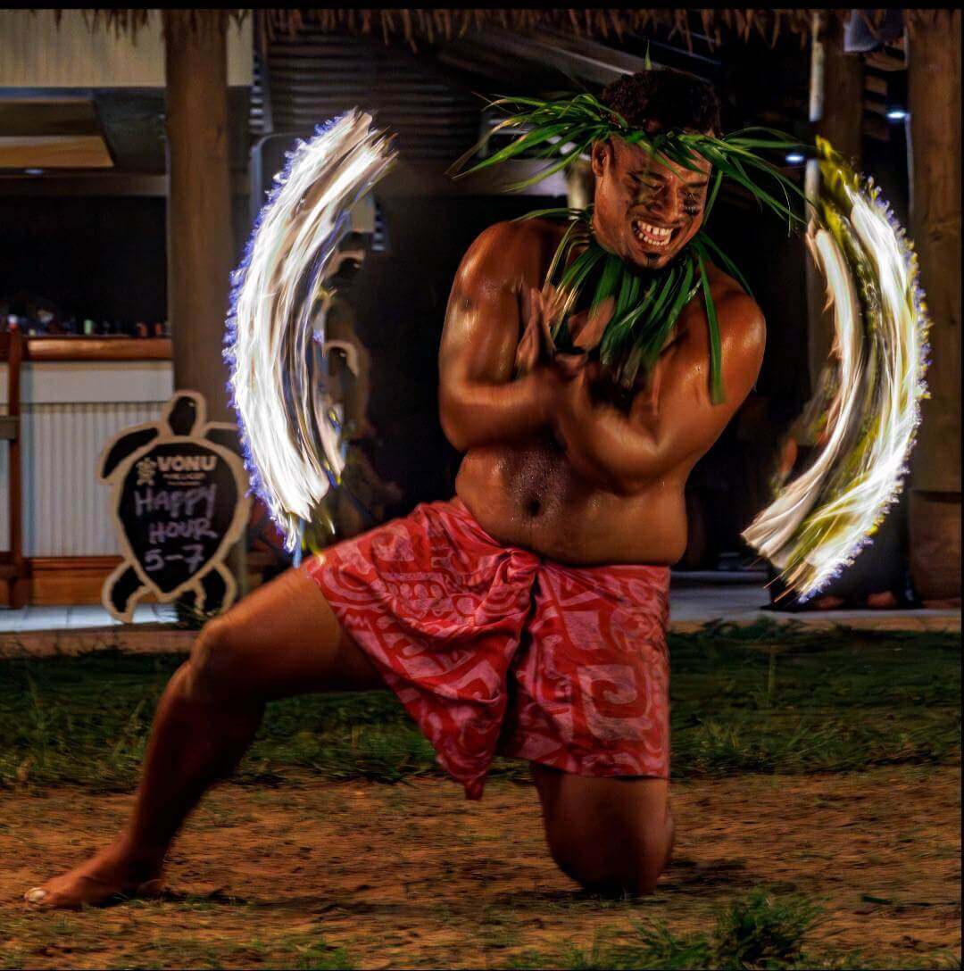 Energetic Fijian fire dancer in traditional attire at Uprising Beach Resort, spinning brilliant arcs of flame at night.