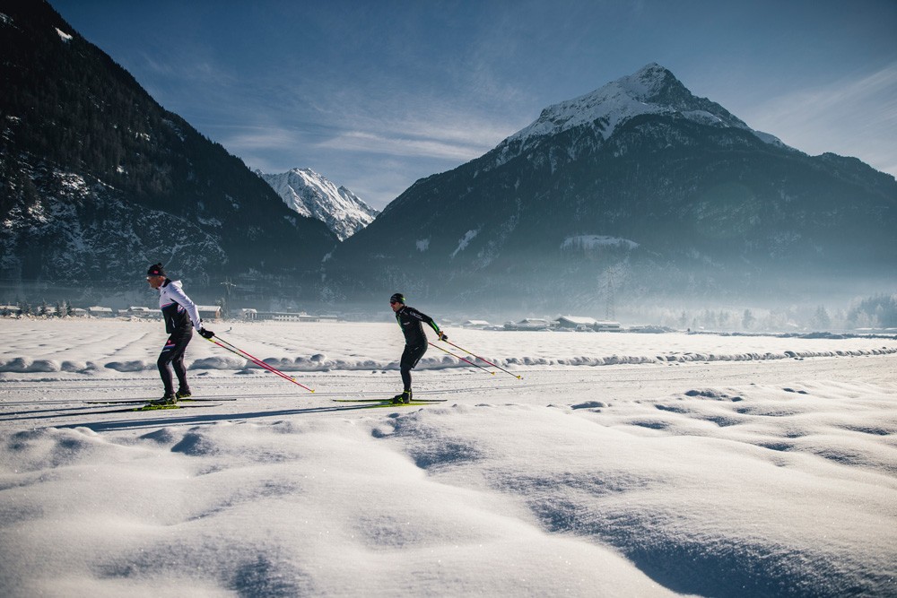 Zwei Personen beim Langlaufen im Ötztal in einer verschneiten Landschaft mit majestätischen Bergen im Hintergrund unter einem klaren blauen Himmel.