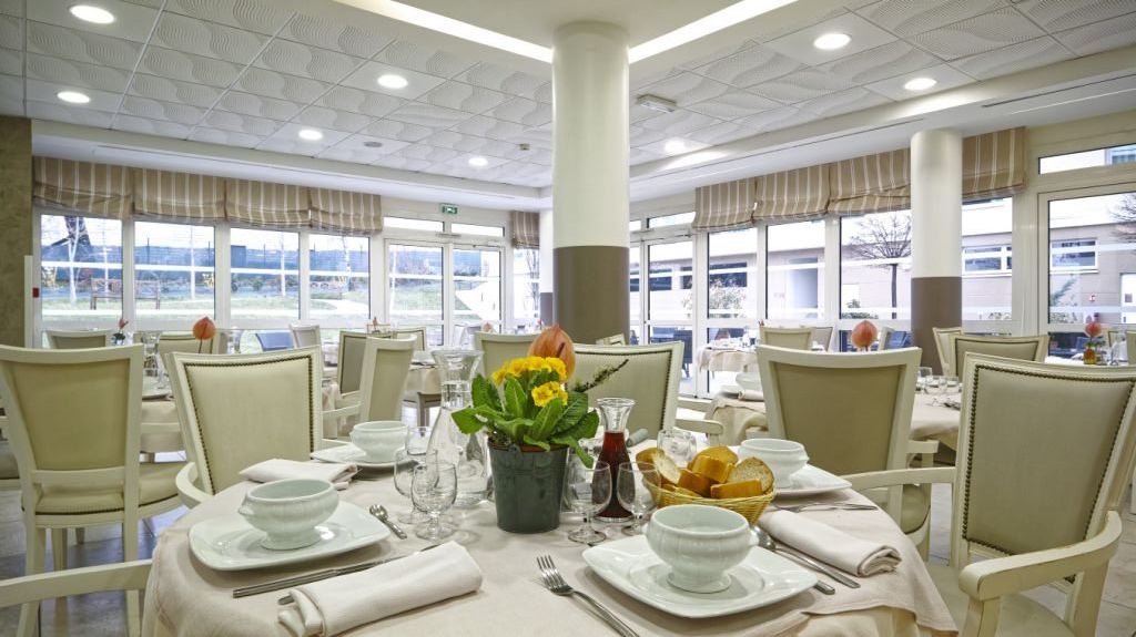 Elegant dining room with cream upholstered chairs, formally set tables with white linens, yellow floral centerpieces, and large windows with striped valances