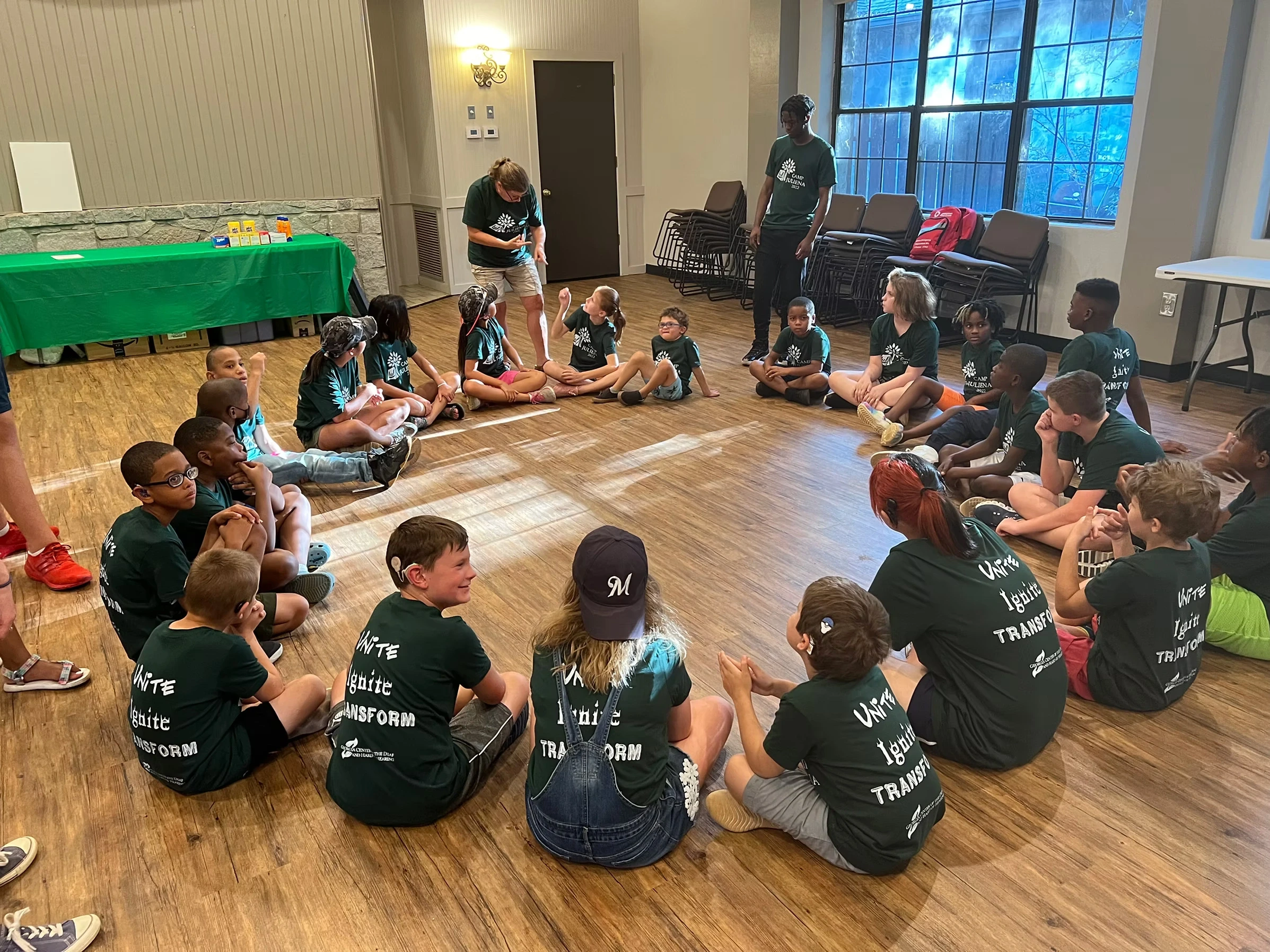 A group of campers and staff sit in a circle on the floor, engaged in an activity. They wear matching green Camp Juliena t-shirts.
