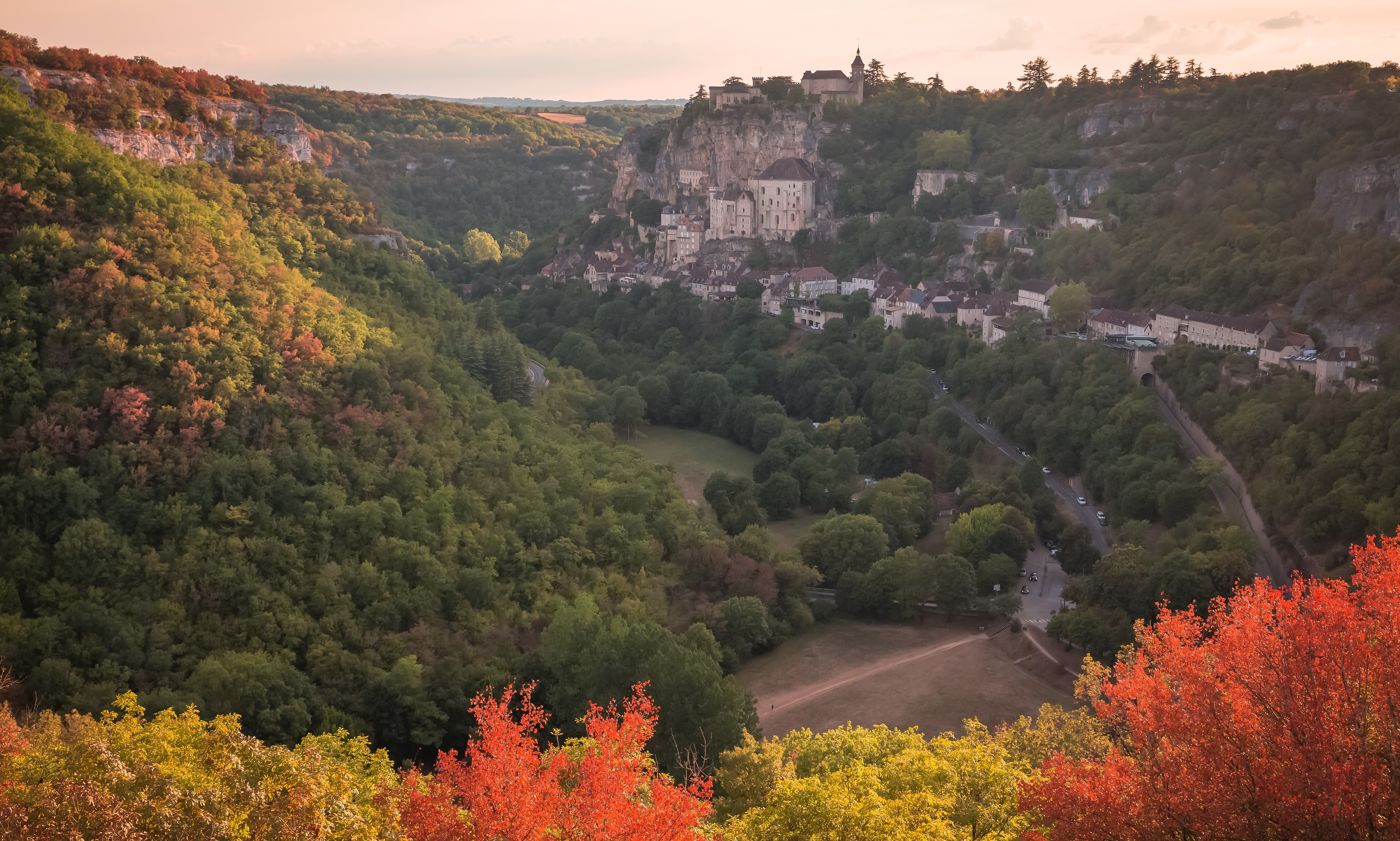 A picturesque hilltop village surrounded by a lush green valley with trees turning red and orange in the soft light of sunset.