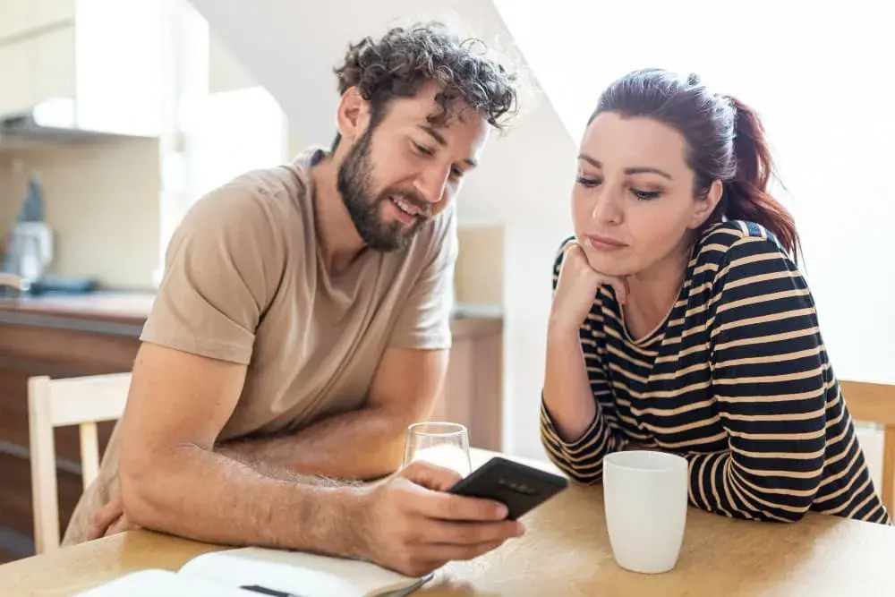 A couple sits at their kitchen table reviewing information on a smartphone, appearing thoughtful and engaged. The warm, natural light reflects a moment of learning and decision-making, representing homeowners exploring refinancing options with Chris Lewis Home Loans.