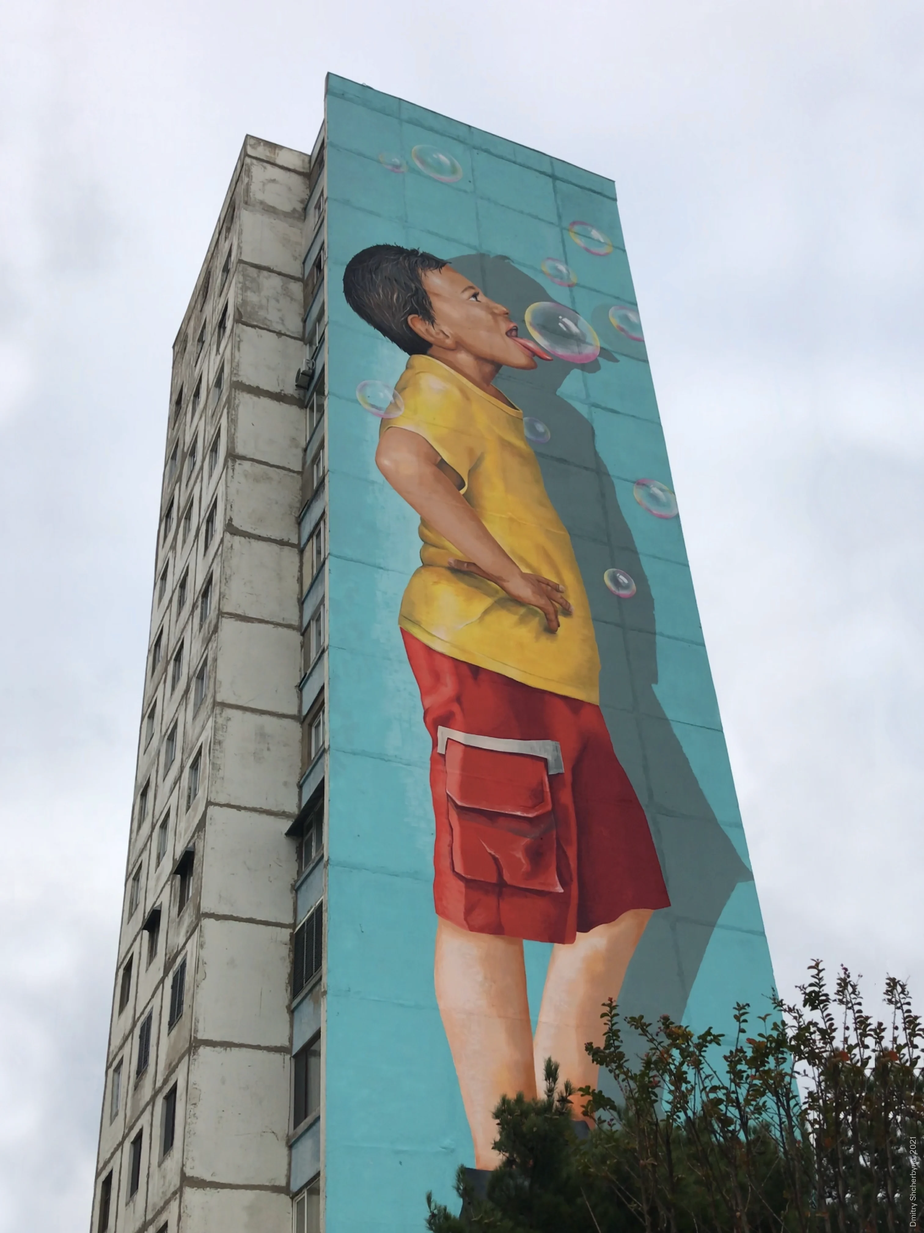 A photorealistic mural on the side of a tower block shows a barefoot boy in a yellow shirt and red cargo shorts standing in profile, blowing soap bubbles against a bright turquoise background.