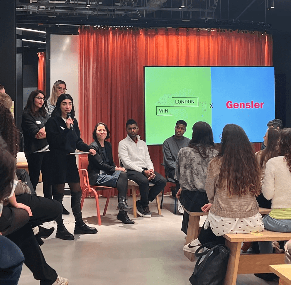 A group of people sits in a discussion setting, with a large screen displaying colorful text in the background.