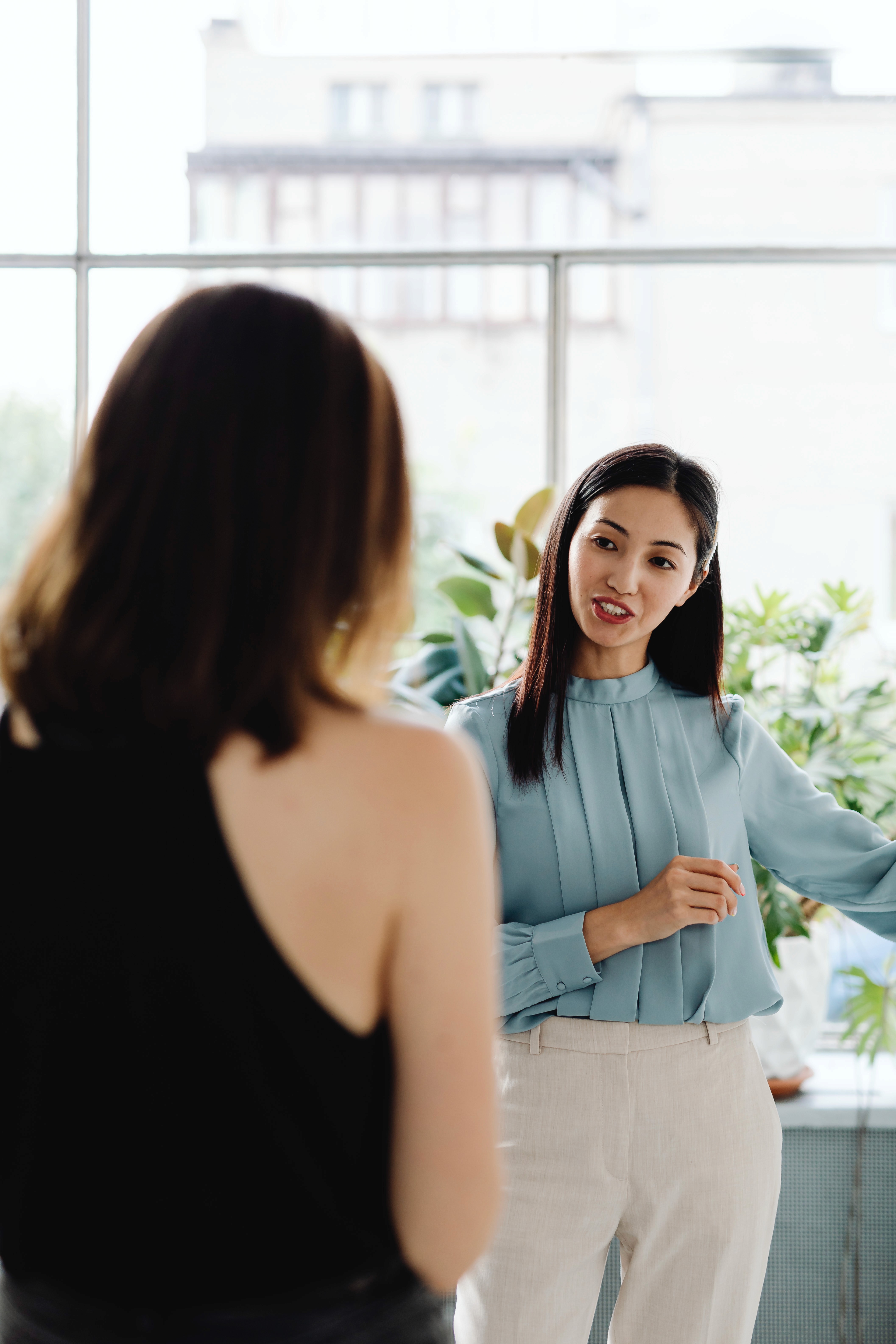 A woman speaks to a woman in a bright office with large windows and plants.