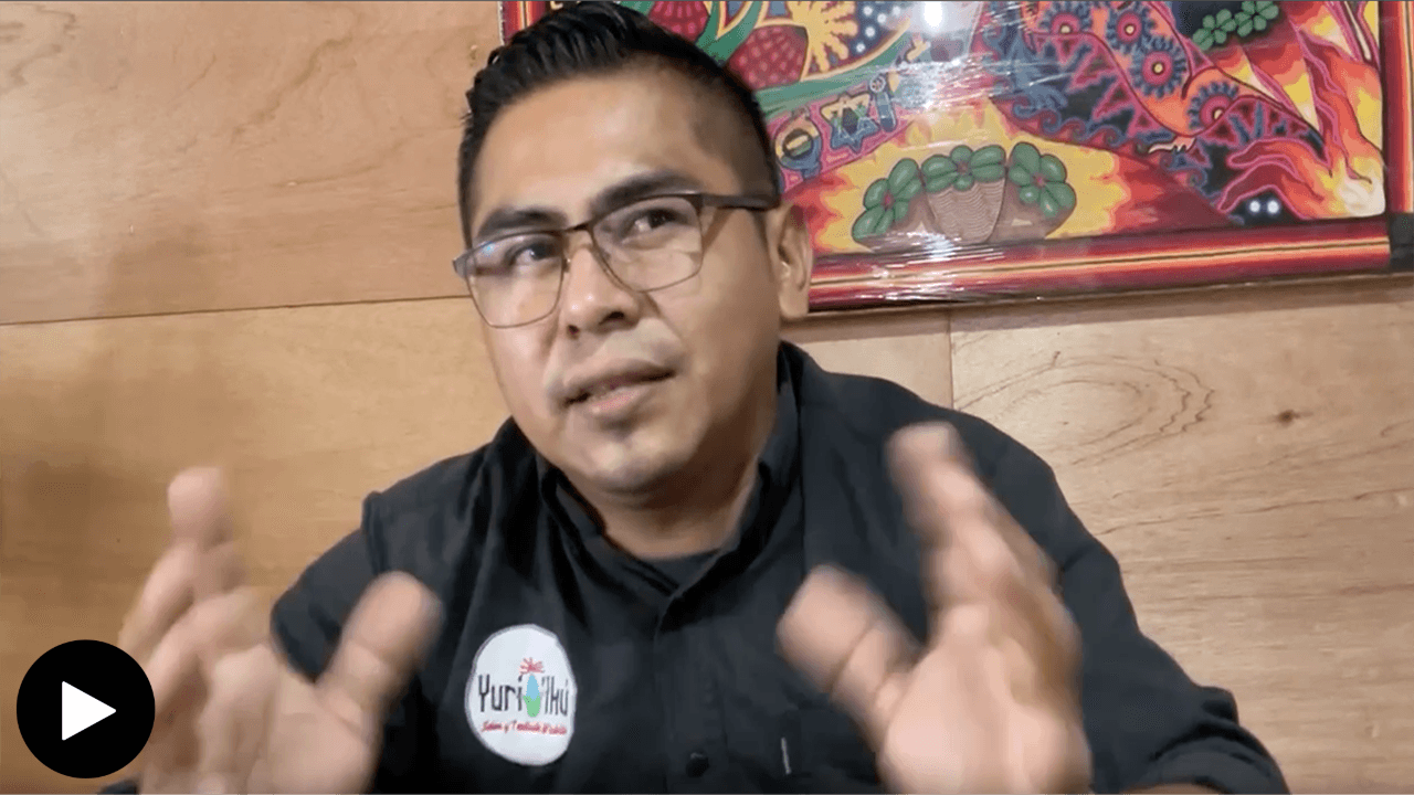 Tutupika Carrillo de la Cruz, actively speaking. His hands are slightly raised forward, his eyes looking toward the speaker on the right. He wears a black shirt and glasses. A multicolor painting sits slightly off-frame behind him.