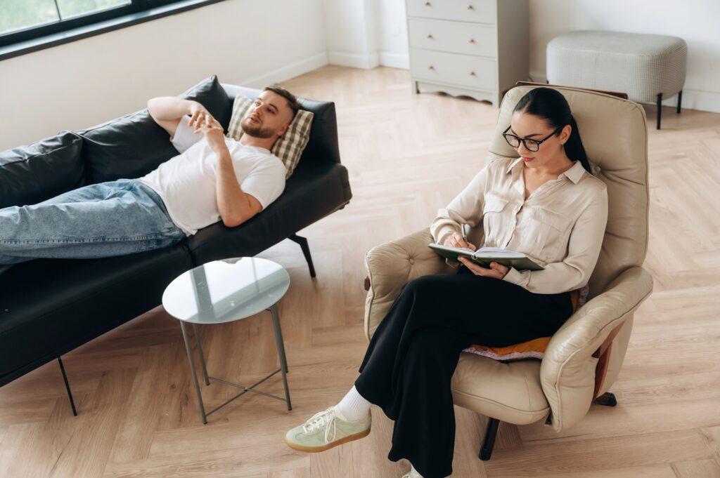 A person lies on a couch while another sits nearby, engaged in a conversation in a modern room.