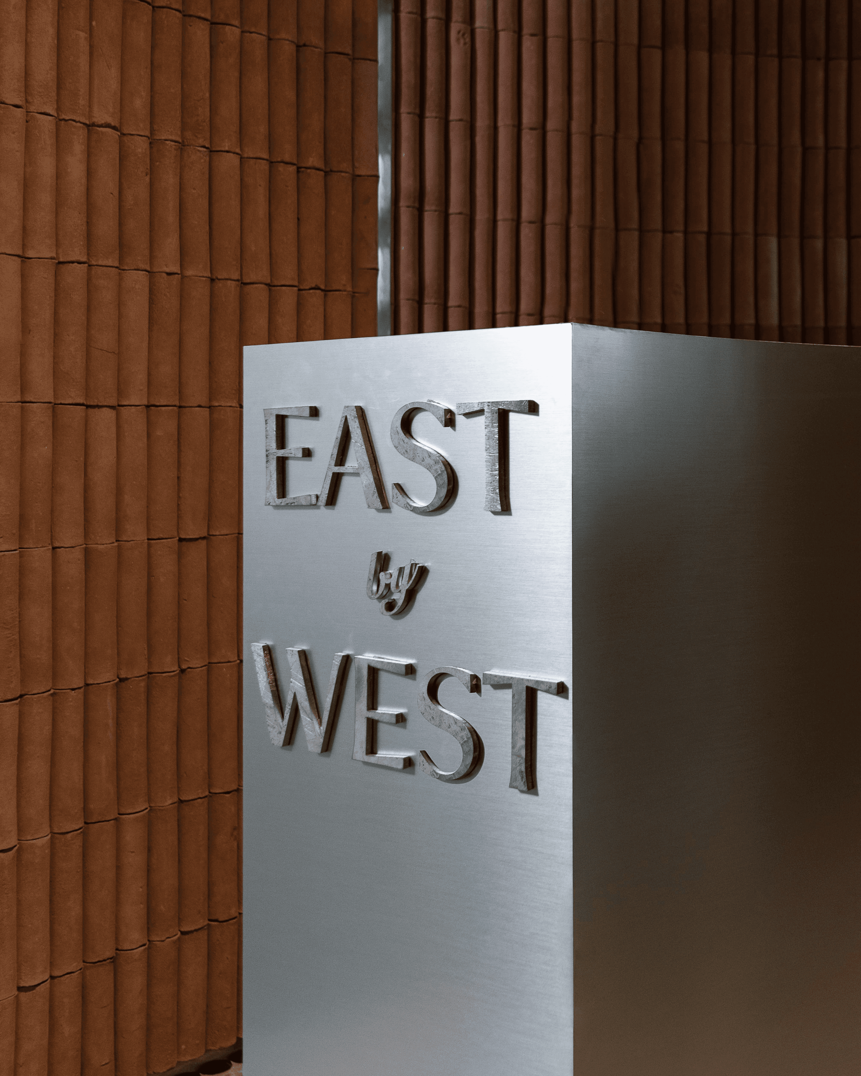 Tall brushed silver pedestal engraved with "East by West" logo against backdrop of warm terracotta brick wall panels.