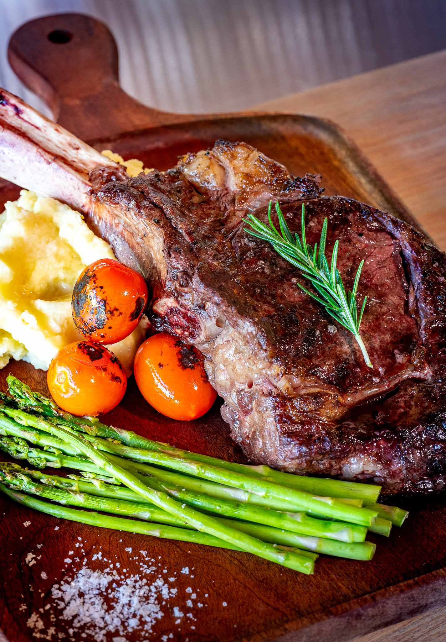 Succulent ribeye steak garnished with rosemary, served with grilled asparagus, cherry tomatoes, and creamy mashed potatoes.