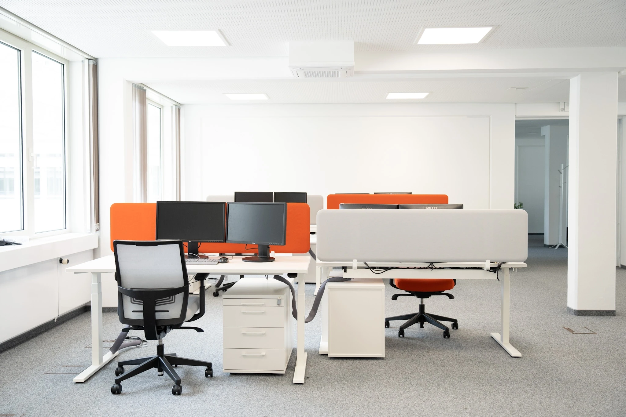 Open-plan office at NFON with white height-adjustable desks, orange and grey acoustic screens, monitors, a grey and an orange office chair and white drawer pedestals on grey carpet.