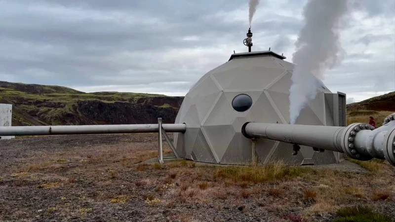A geodesic dome-shaped geothermal wellhead structure in a rugged, grassy landscape under a cloudy sky. Large metal pipes extend horizontally from the structure, and white steam is venting from a stack on top and from the side.