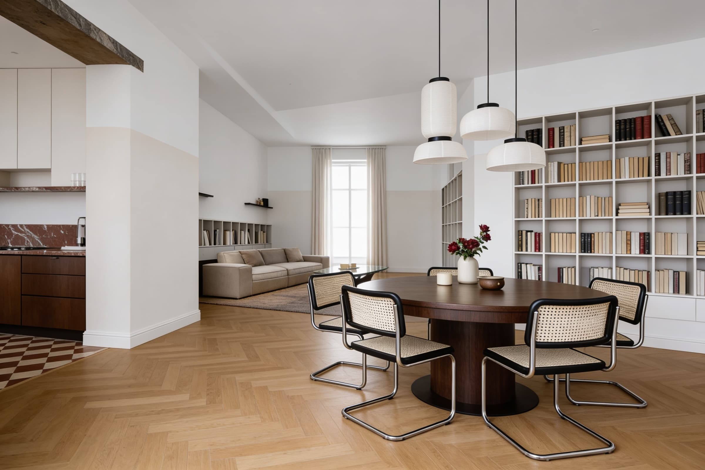 A modern, open-concept dining area features a round wooden table with four sleek, metal-framed chairs, illuminated by three pendant lights, against a backdrop of a cozy living room and a minimalist kitchen.