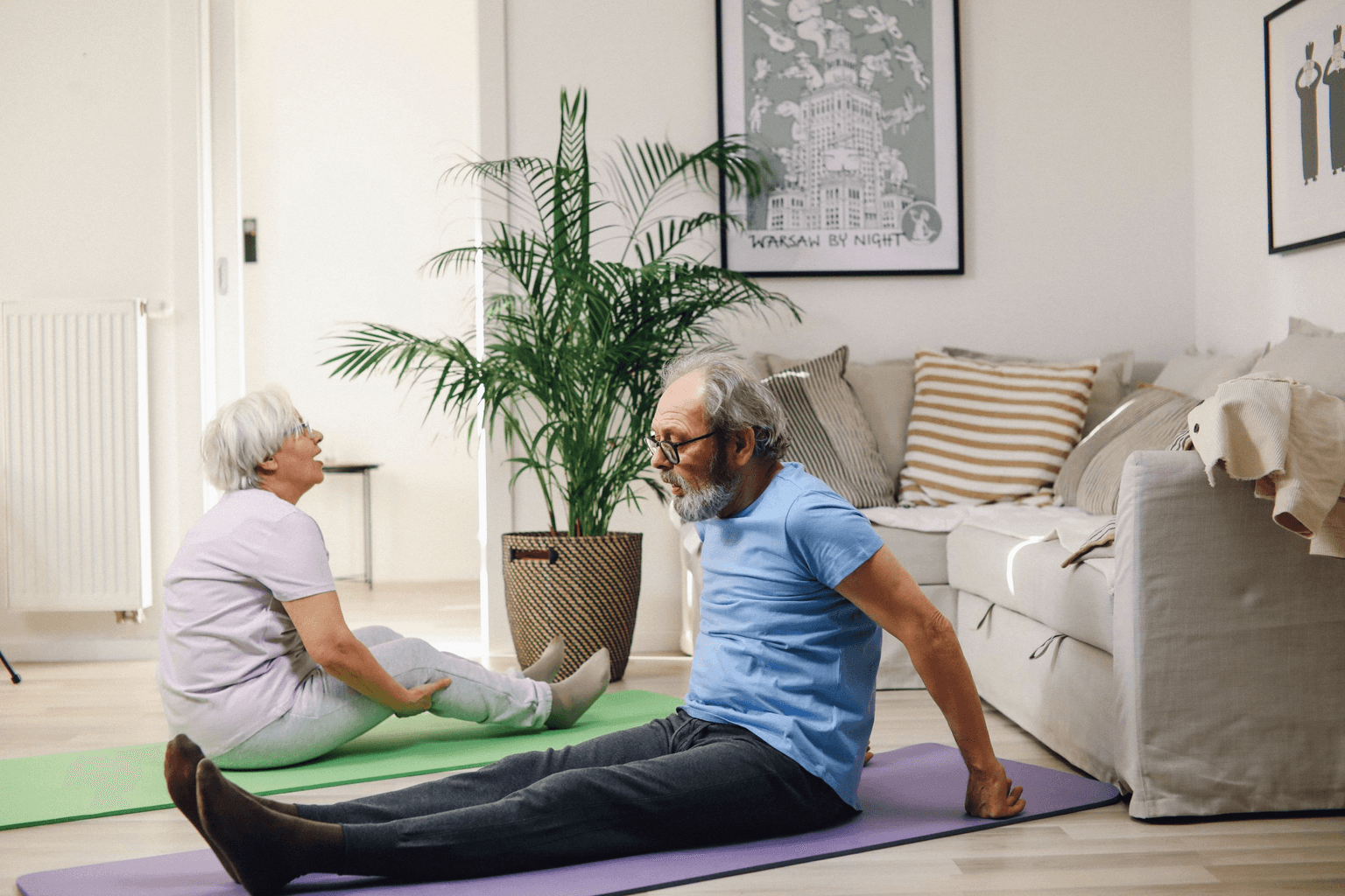 An older couple sitting on exercise mats in their living room, stretching and talking as they go.