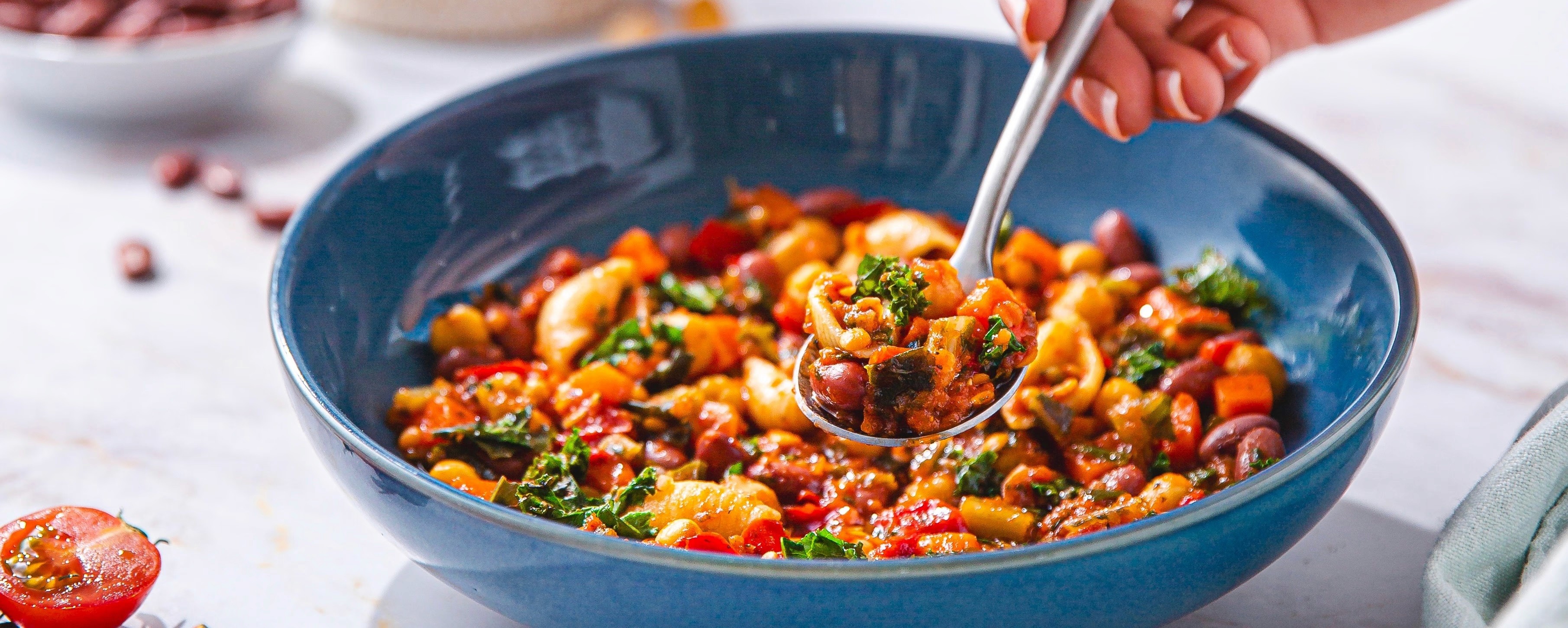 Close-up of a spoon lifting a hearty vegetable and bean stew from a blue bowl.