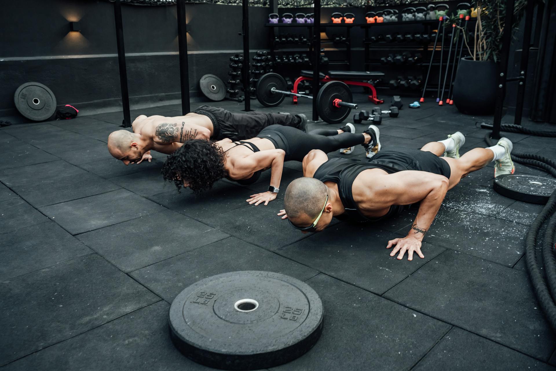 A group of people with dark fitness attire perform a push-up on the dark gym floor.