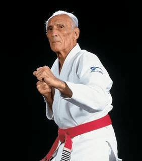 Elderly Gracie family member in white gi and red belt demonstrating a move