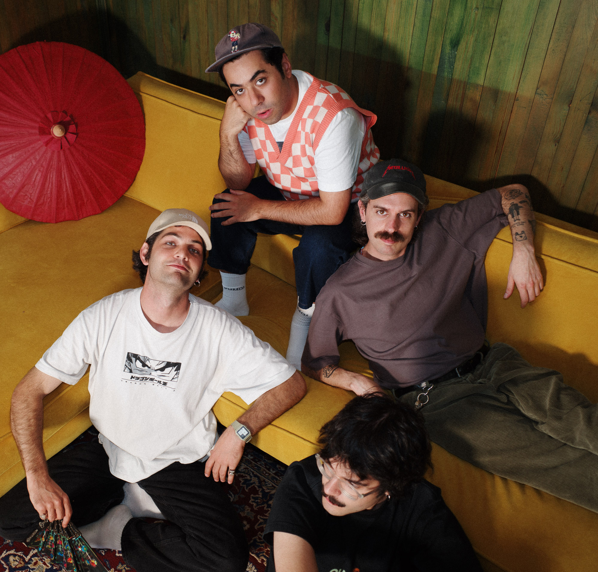 A group of four people casually pose in a cozy room featuring a yellow sectional sofa, while a red paper parasol leans against wood-paneled walls, creating a relaxed and retro atmosphere.