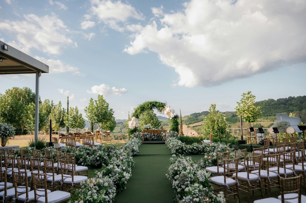 Outdoor hilltop wedding ceremony setup with a flower-lined green aisle, gold chiavari chairs, and a circular greenery arch overlooking a countryside vineyard