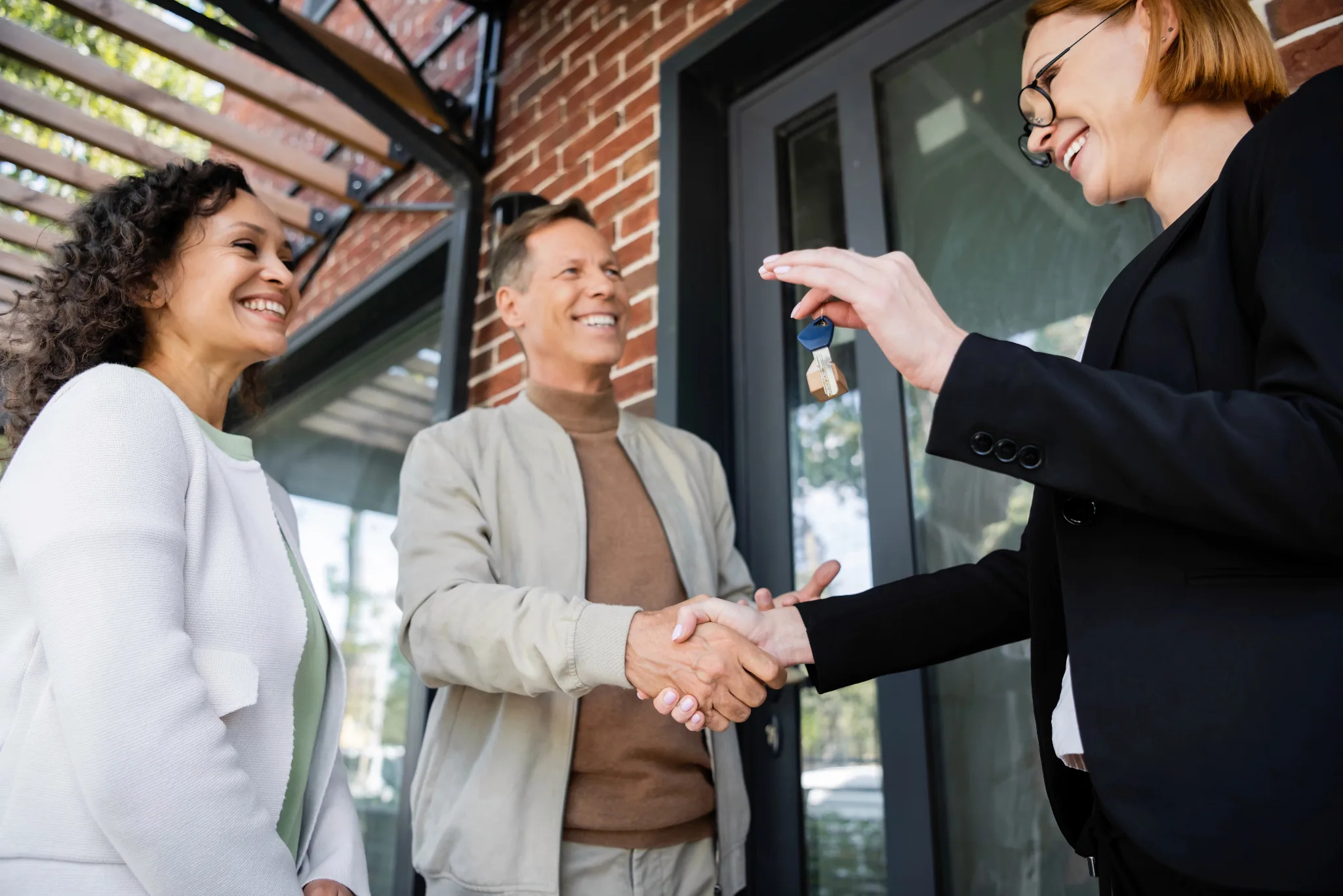 A real estate agent in professional attire hands over house keys to a smiling couple outside a modern brick home with large windows.