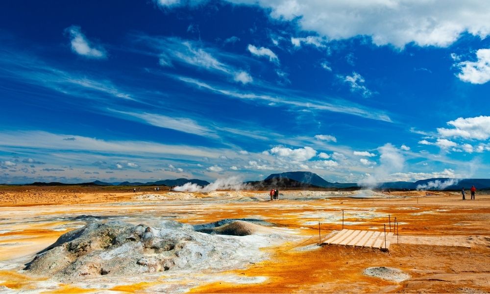 Wide view of the geothermal fields at Námaskarð Pass near Lake Mývatn in Iceland, featuring steaming vents, orange and white mineral-stained earth, walking paths, and distant volcanic mountains beneath a dramatic blue sky.