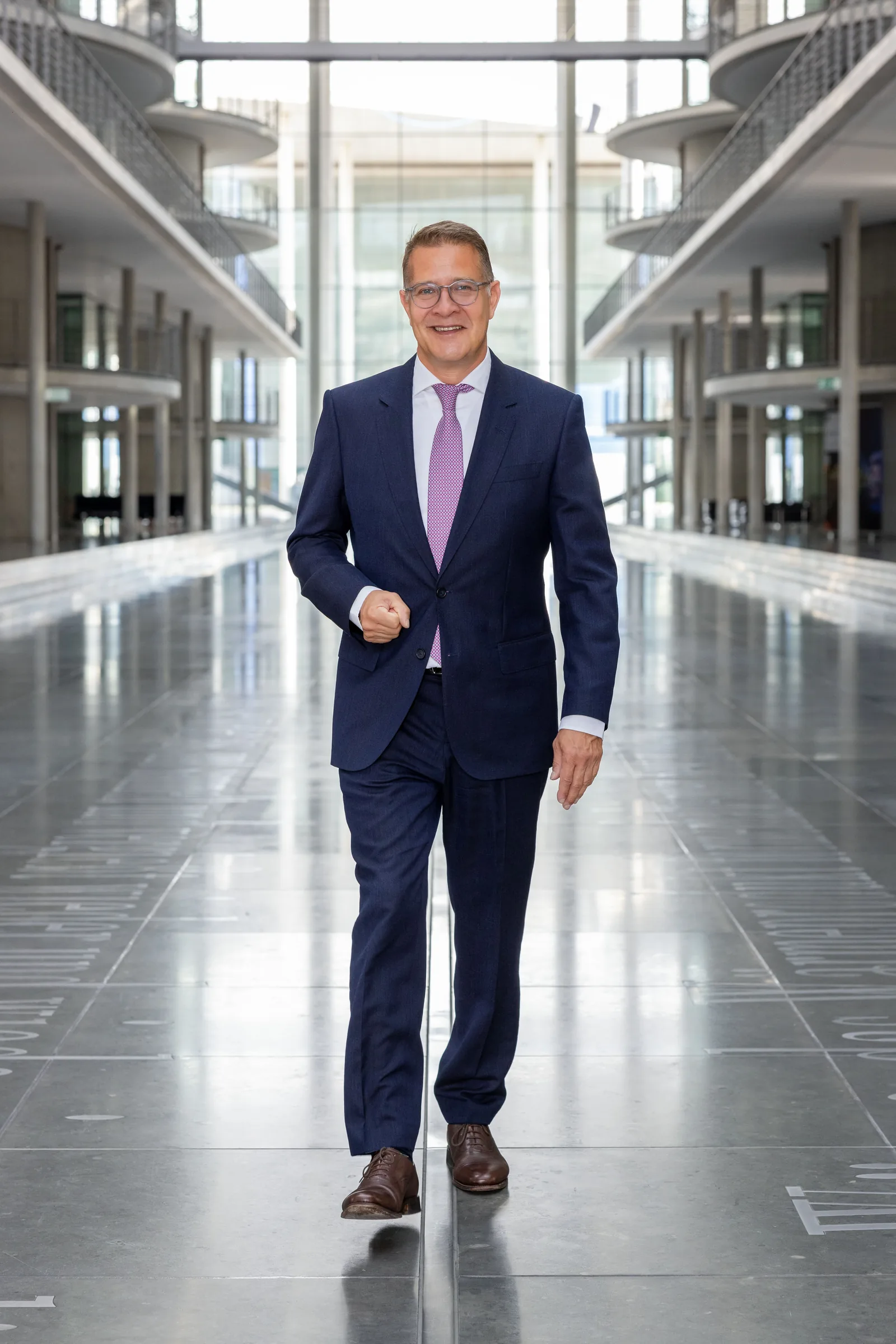 Adrian Grasse geht im Anzug lächelnd durch die moderne Architektur des Paul-Löbe-Hauses im Berliner Regierungsviertel.
