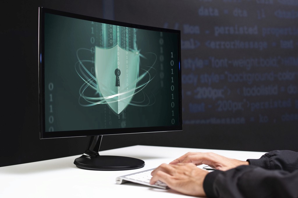 Profissional de TI digitando em teclado diante de monitor exibindo escudo de segurança digital com cadeado e código binário, representando cibersegurança e proteção de dados - FindUP - Antivirus gratuito