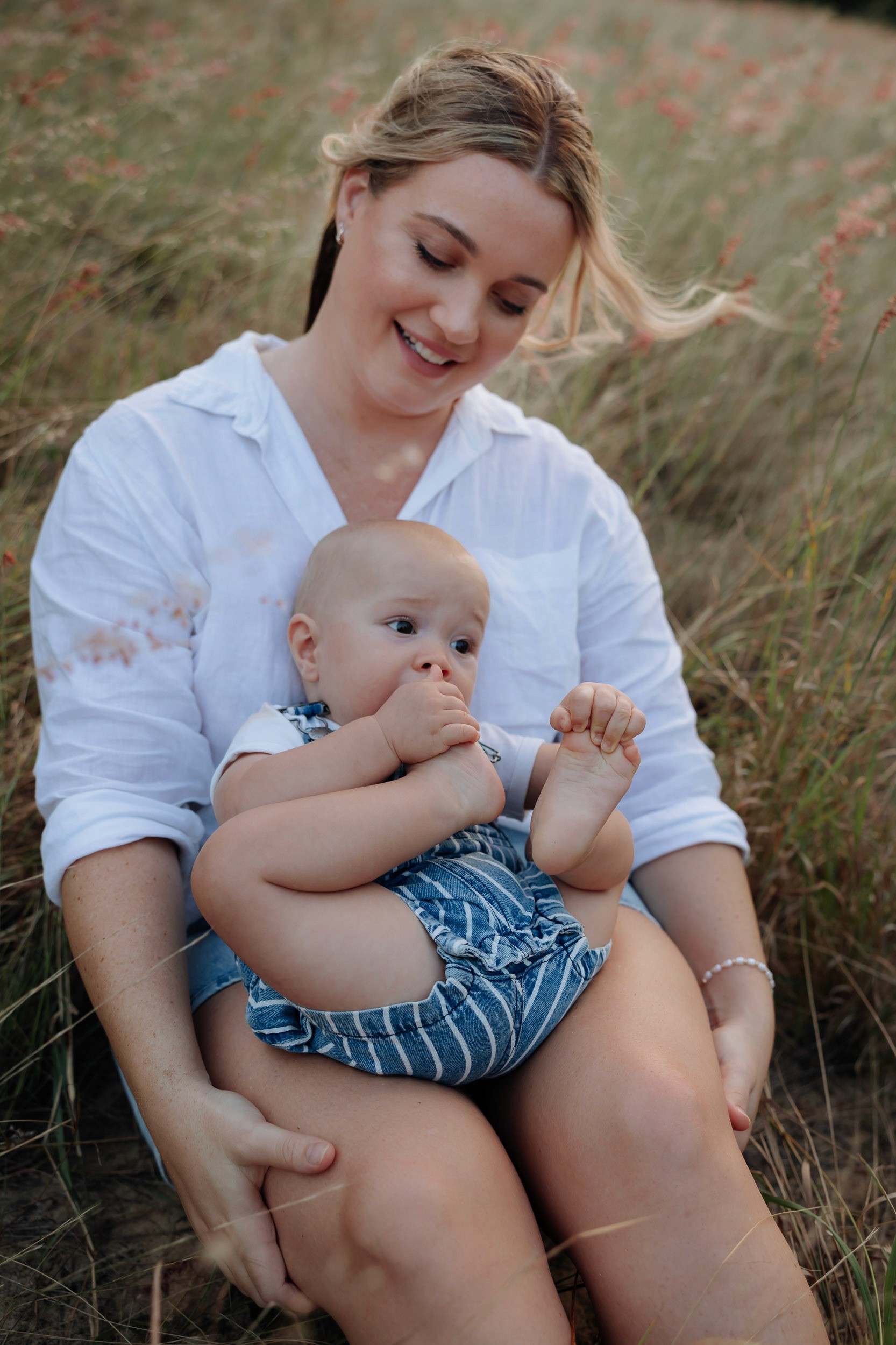 Natural lifestyle image of mother sitting in tall grass with her baby, relaxed outdoor family session in Mackay QLD