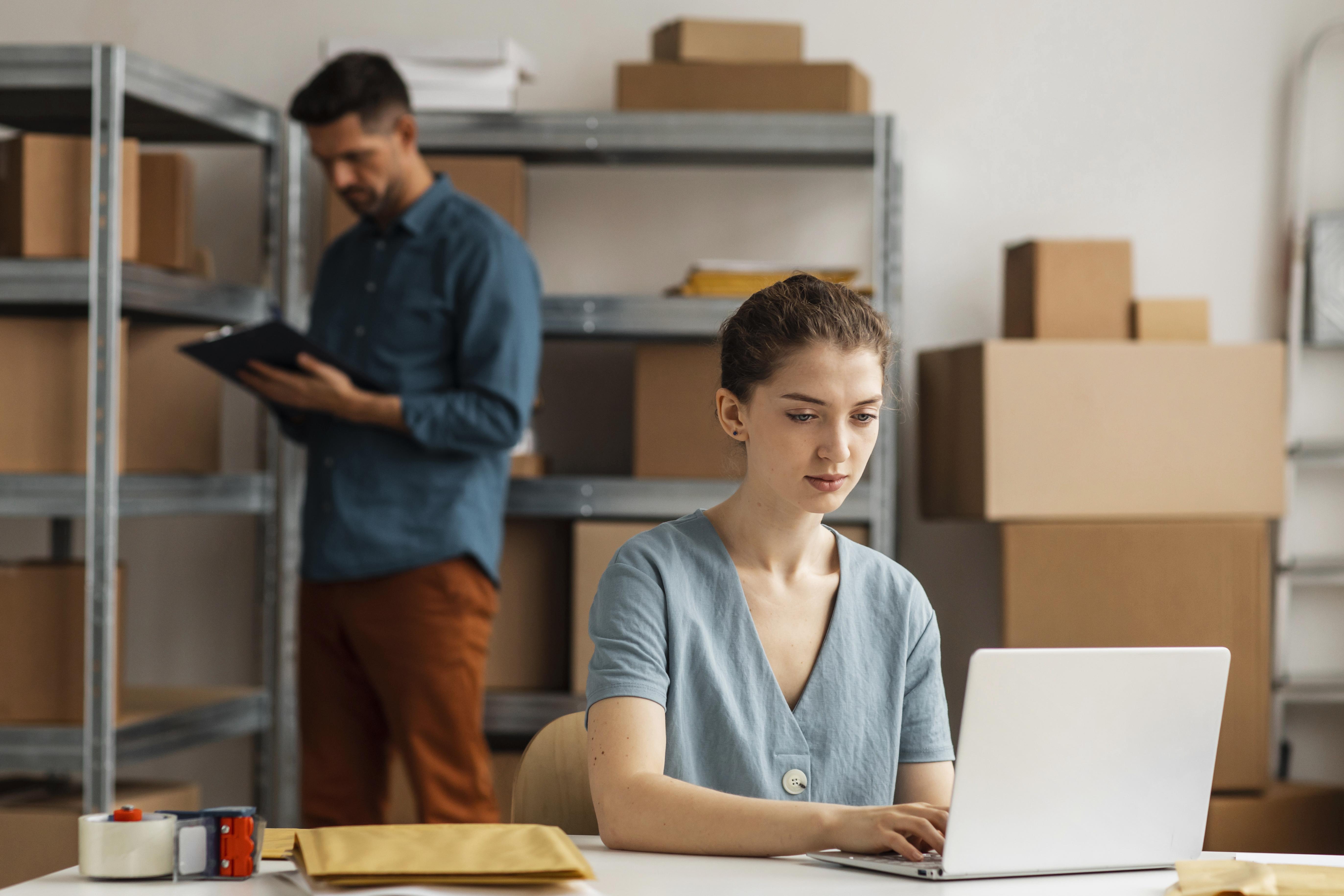Woman focused on a laptop in a warehouse office, surrounded by boxes. A man in the background takes notes, creating a busy yet calm work environment.