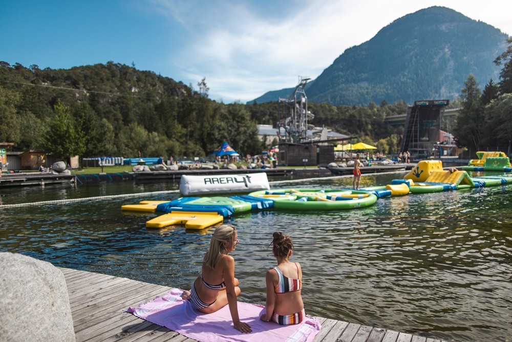 Frauen entspannen sich auf einem Steg am Seeufer in der AREA 47, umgeben von bunten Aufblasbaren spring Elementen im Wasser.