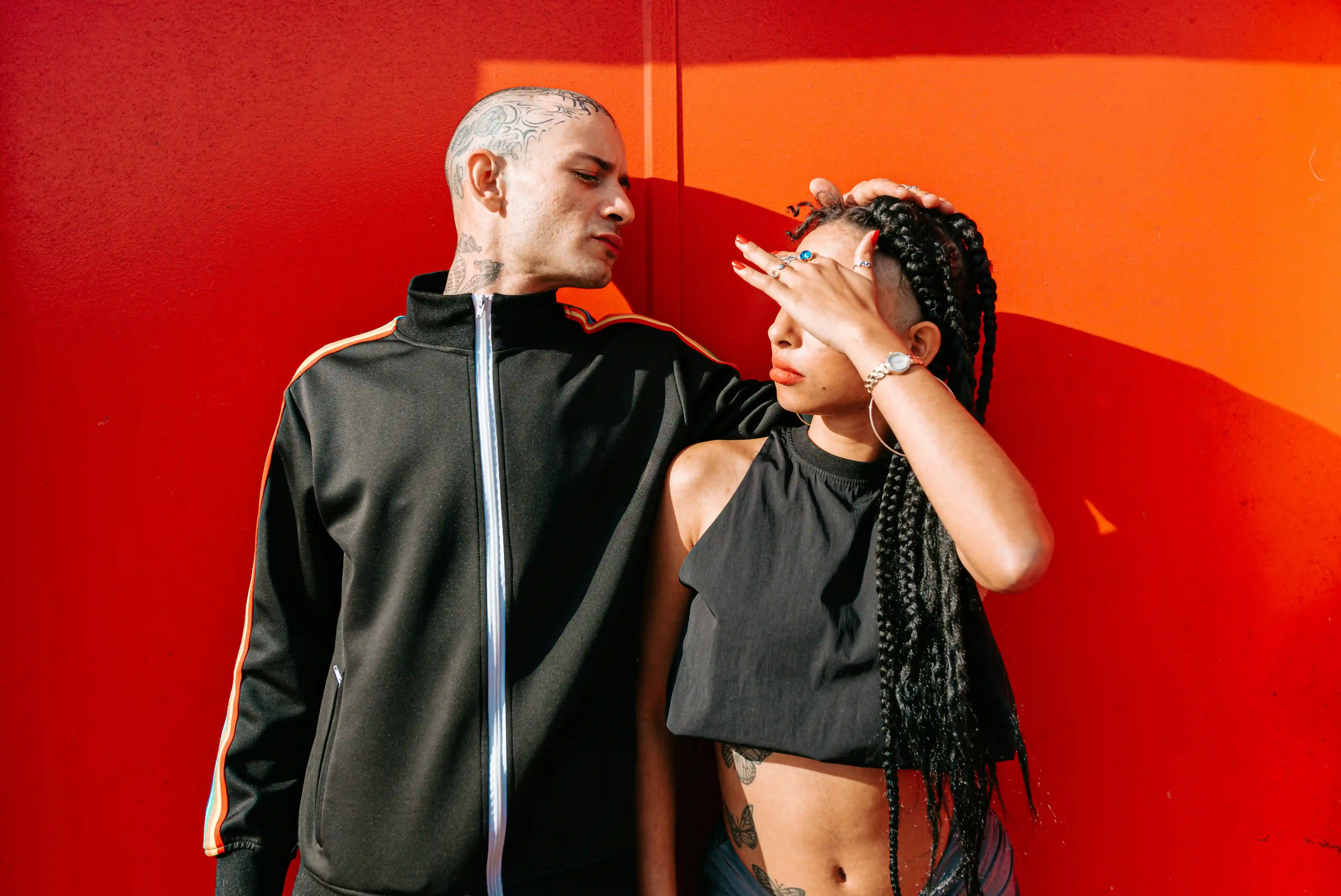 man and woman wearing black tracksuit and crop top stand facing each other against an orange wall