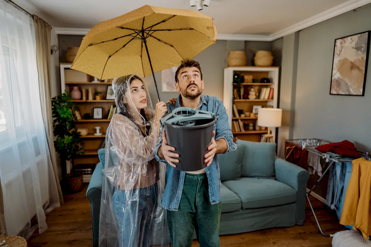 Paar steht im Wohnzimmer mit Schirm, Regencape und Eimer, da es von der Decke tropft