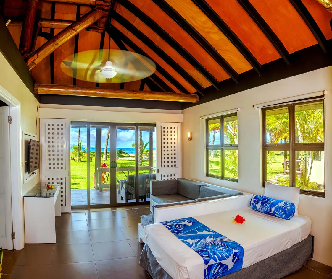 Uprising Beach Resort room with bed, sofa, vaulted ceiling, and sliding glass doors revealing a tropical lawn and the ocean.