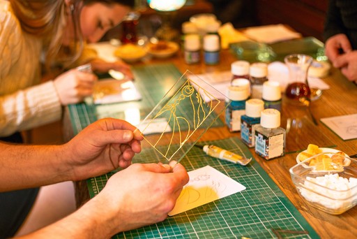 A person crafting a Turkish mosaic lamp, carefully applying multicolored beads to the ornament. The background shows a workshop setting with tools hanging on the wall.