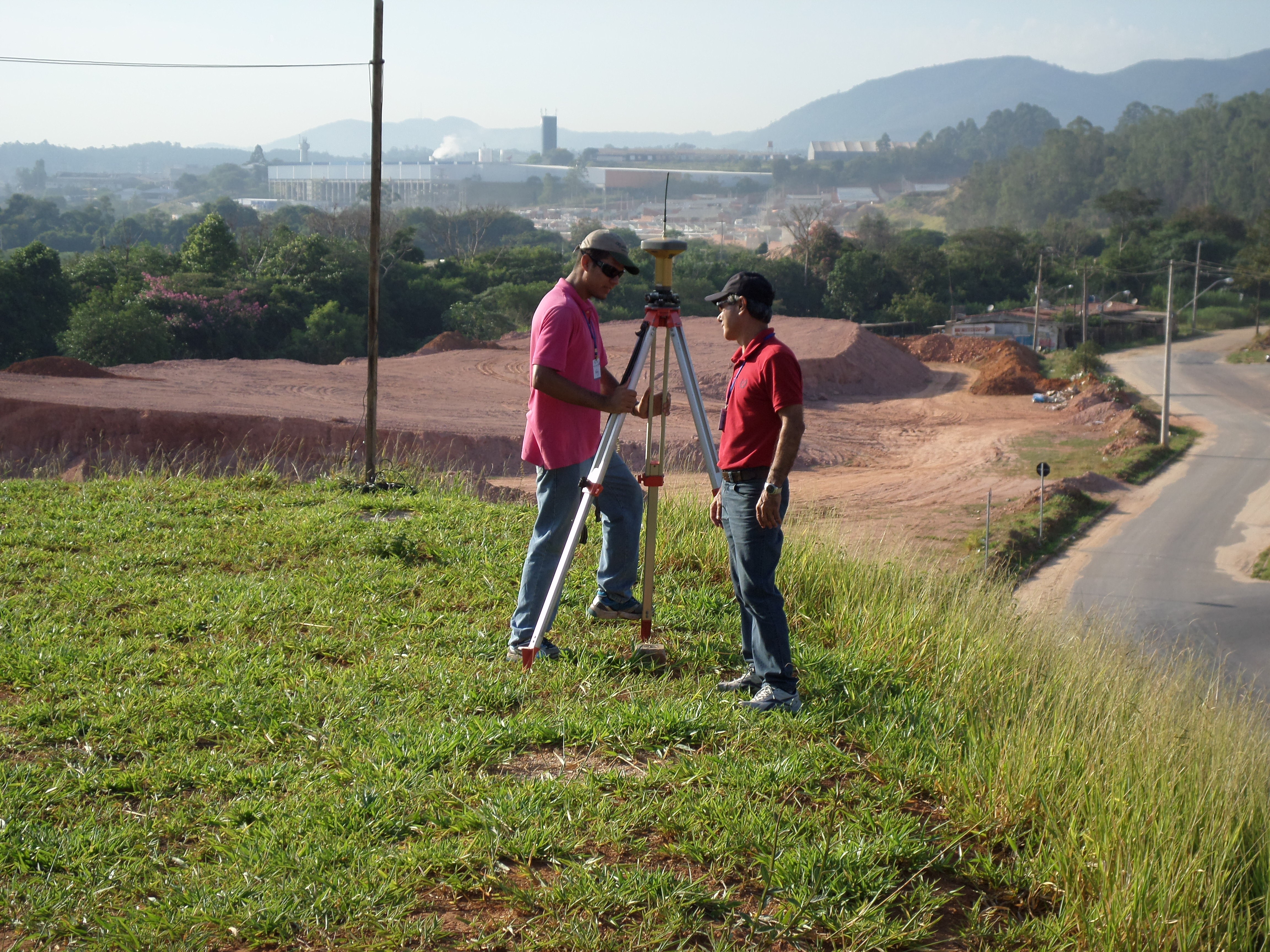 Equipe de topografia operando uma estação total sobre tripé em um terreno elevado com vista para uma estrada e obras de infraestrutura ao fundo.