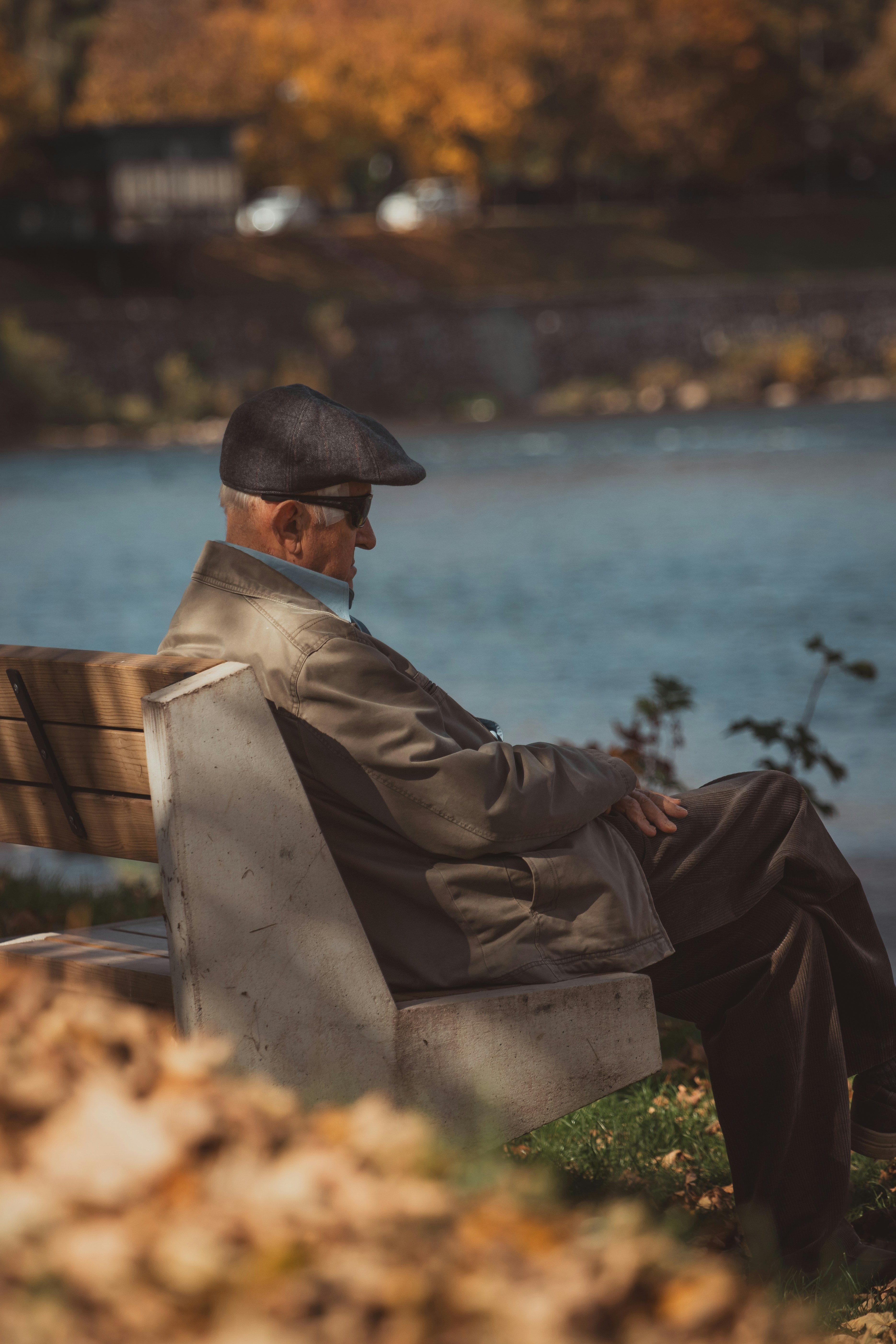 Elderly man in cap sitting on park bench by water.