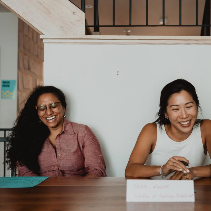 Two women sitting at a table, smiling and laughing. One wears glasses and a maroon shirt, the other a white sleeveless top. Bright, cheerful ambiance.