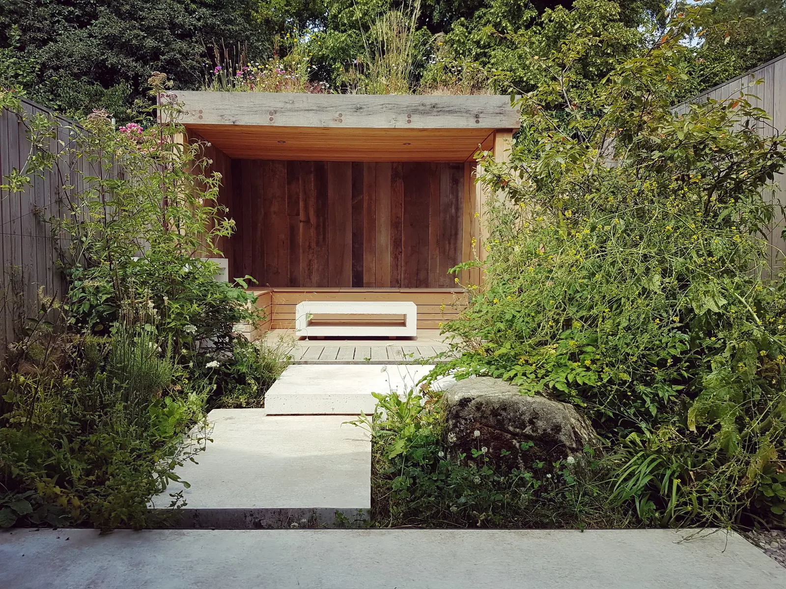 A pathway leads to a wooden doorway, framed by lush greenery and plants on either side.