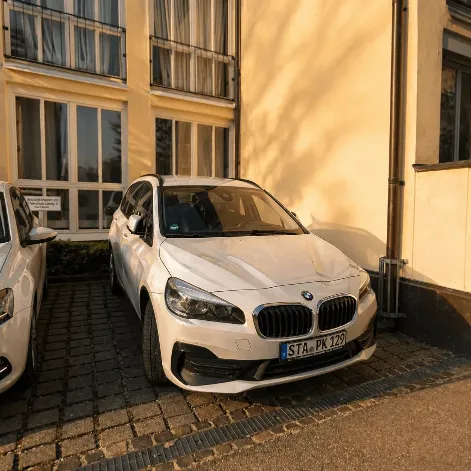 Ein weißer BMW 2er Gran Tourer als Fahrschulwagen mit dem Kennzeichen STA-PK 129 parkt in der Einfahrt vor einem gelben Gebäude.
