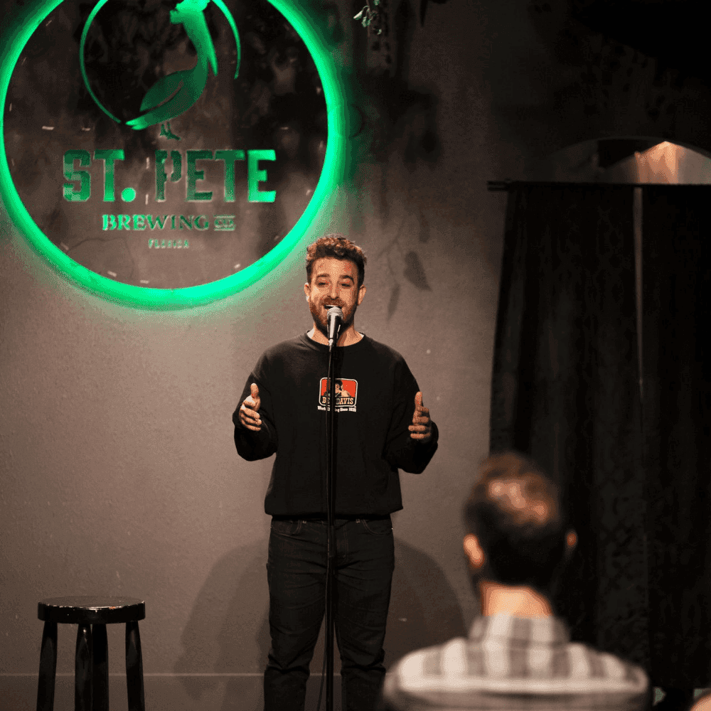 A male comedian making a hand gesture on stage at a St Pete Standup show