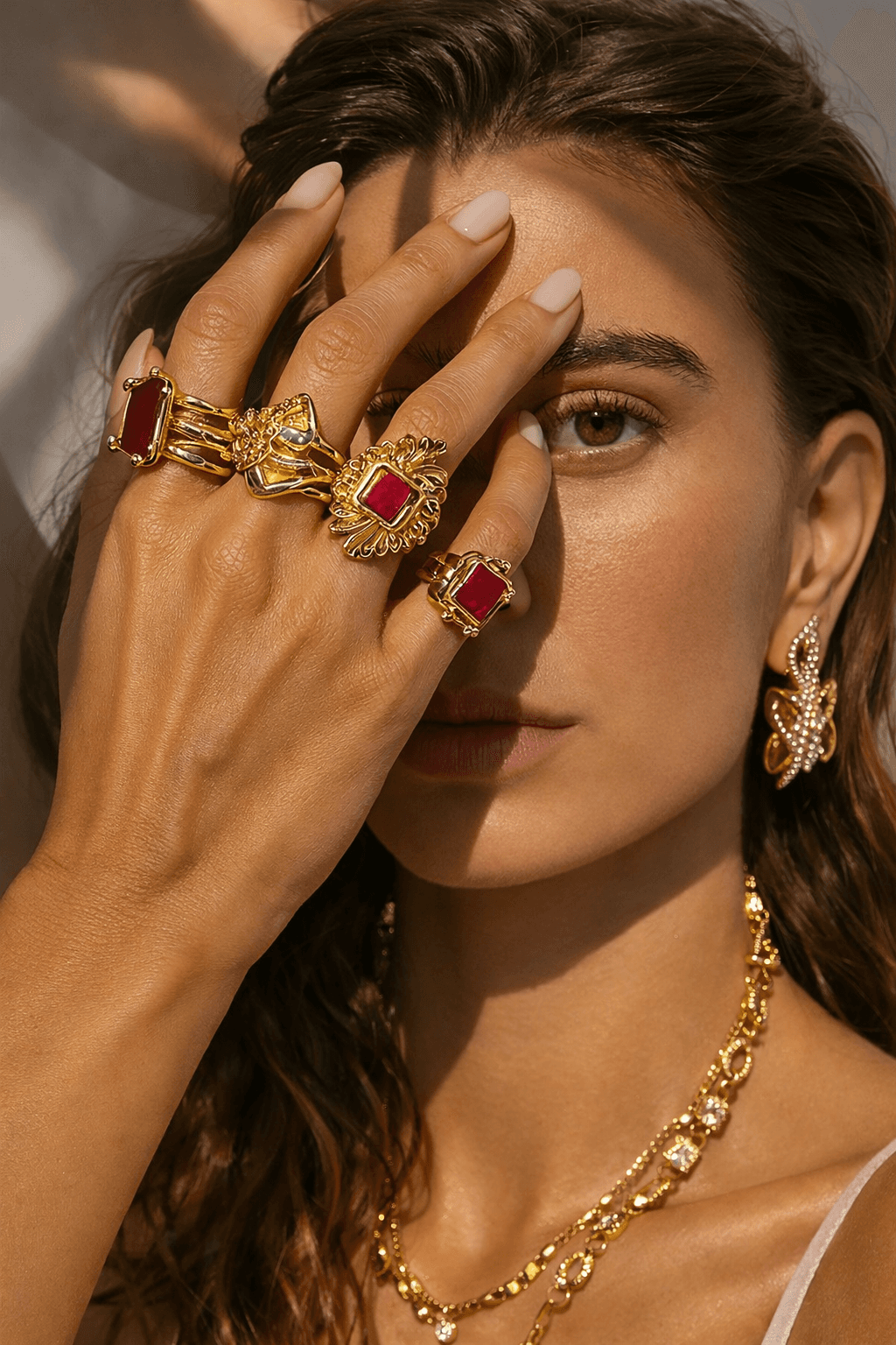 Close-up of a woman showcasing ornate gold jewelry, including chunky rings with red gemstones, a layered necklace, and butterfly earrings, under warm, shadowed lighting.
