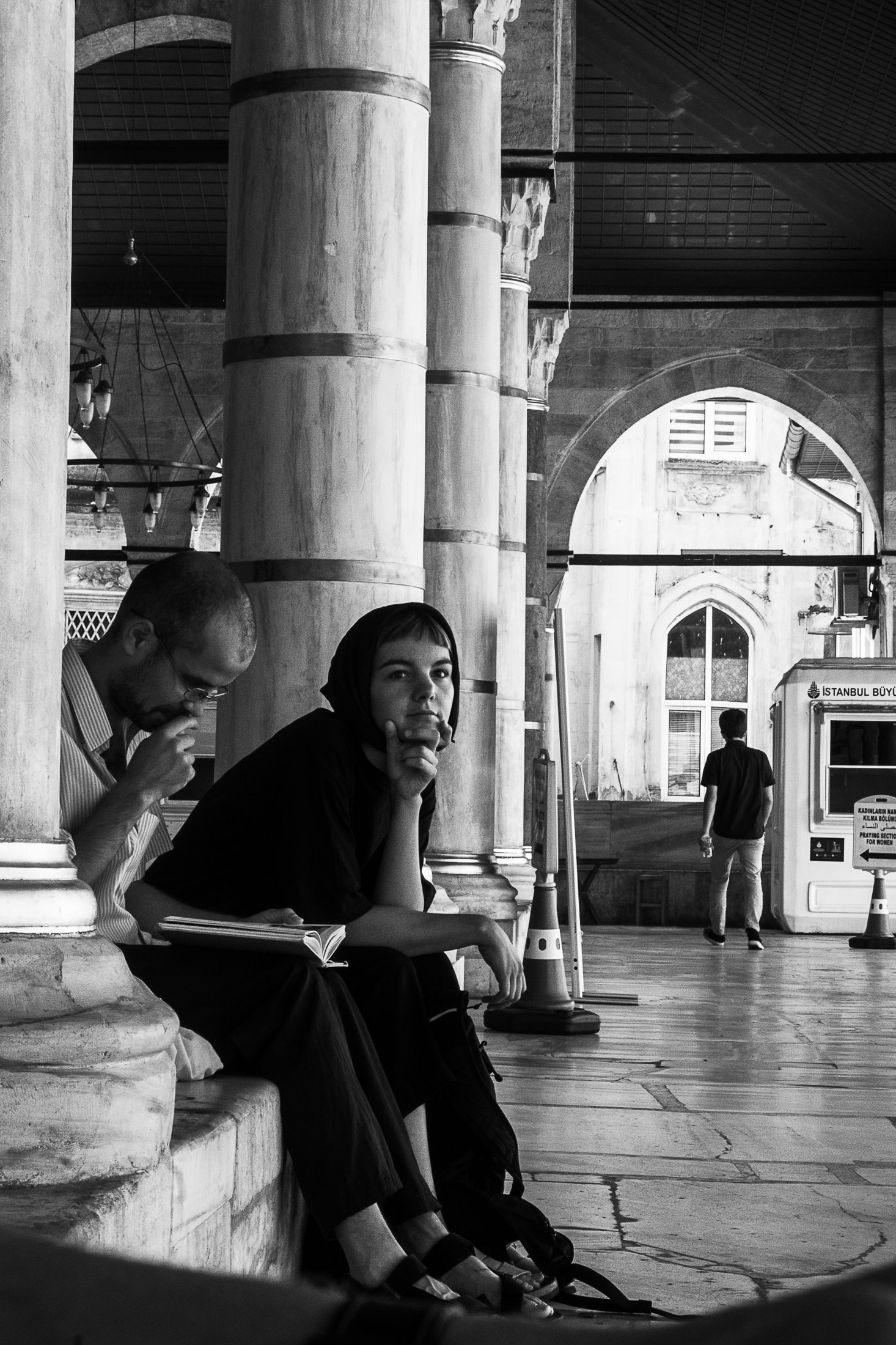 Woman sitting beside marble columns looking toward the camera, man reading next to her, arched architecture behind.