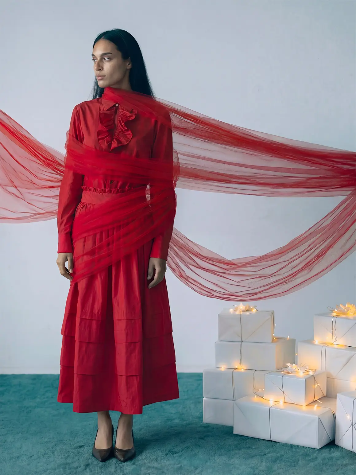 A model in an elegant red dress stands beside a series of wrapped white gift boxes with glowing lights, set against a minimalist backdrop decorated with draped red tulle, embodying high-end visuals for an AI production project in a fashion brand context.