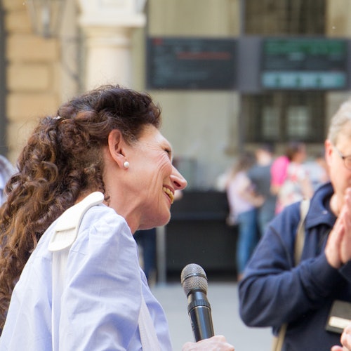 A woman holding a microphone smiles at another person in an outdoor setting, with blurred people and buildings in the background.