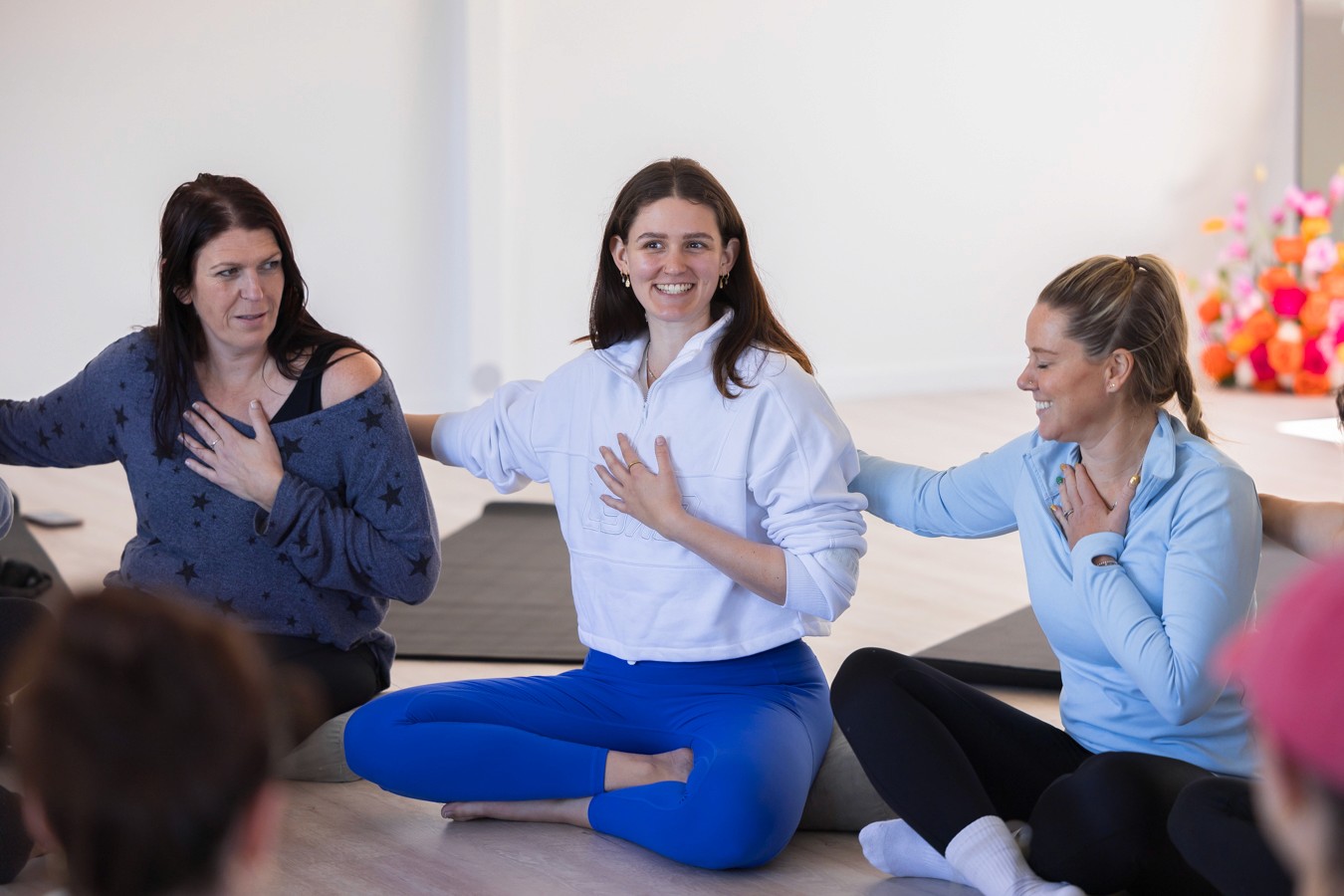 Women sitting on floor cross legged with left hands on heart and right hands on each other's shoulder