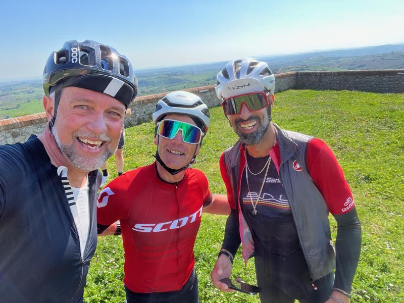 Three cyclists in colorful gear smile and celebrate atop a grassy hill with a scenic landscape in the background on a clear, sunny day.