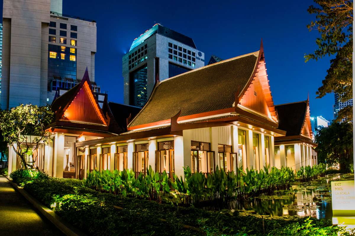 A traditional Thai-style building with a tiered orange roof and lit interior sits in front of modern skyscrapers at dusk.