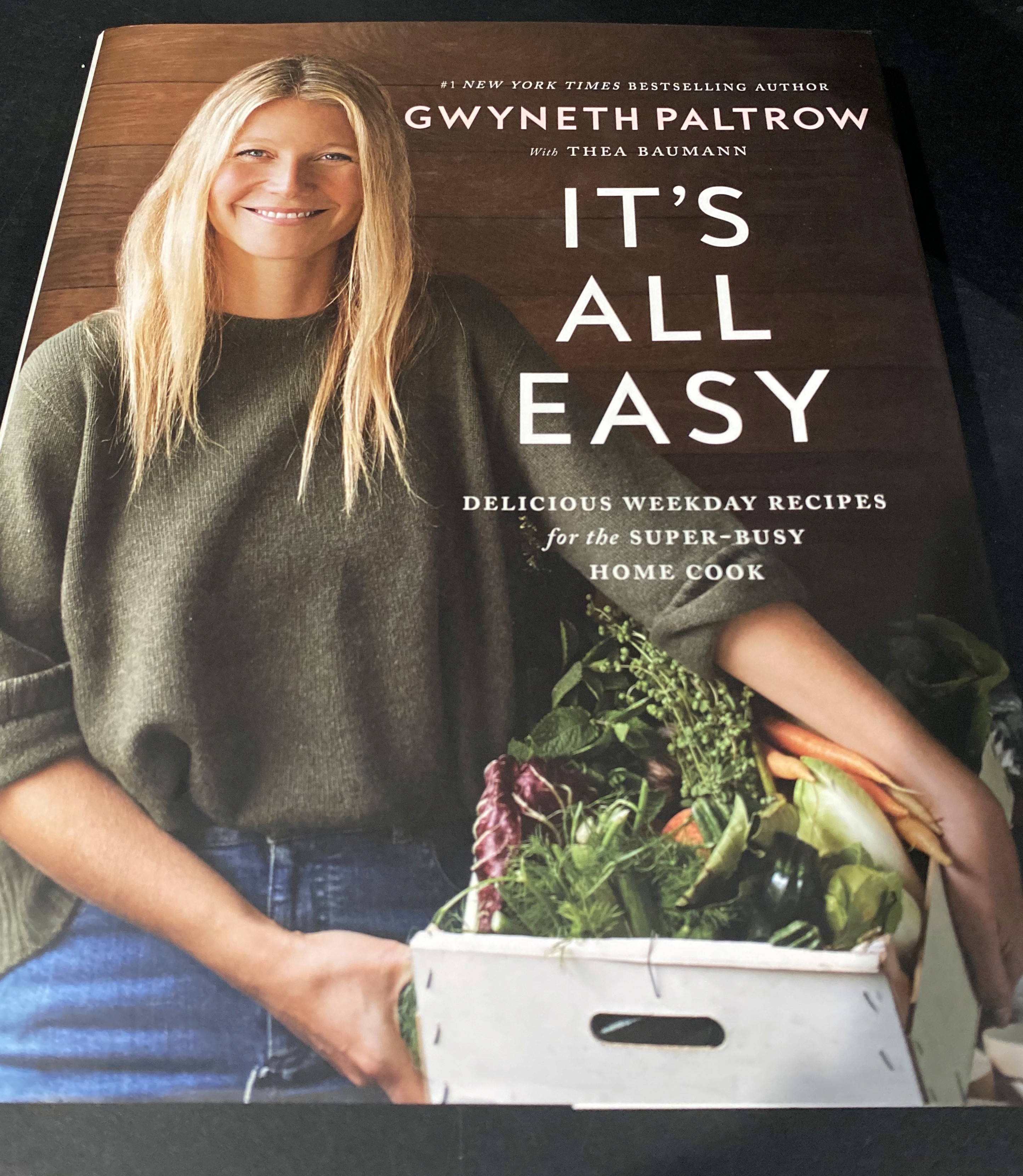 A cookbook titled "It's All Easy" by Gwyneth Paltrow is displayed, showcasing a cover image of a person holding a box of fresh vegetables, with a focus on providing delicious recipes for the super-busy home cook.