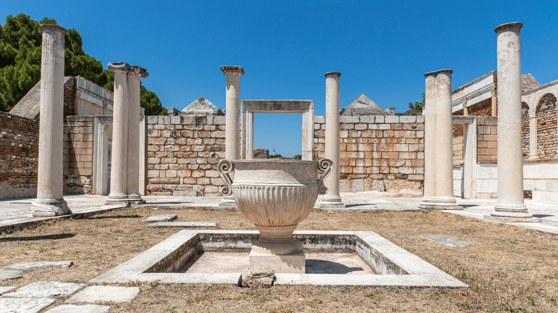 Ruins of the ancient synagogue in Sardis, Türkiye — Marble columns and a large stone urn under a bright blue sky.